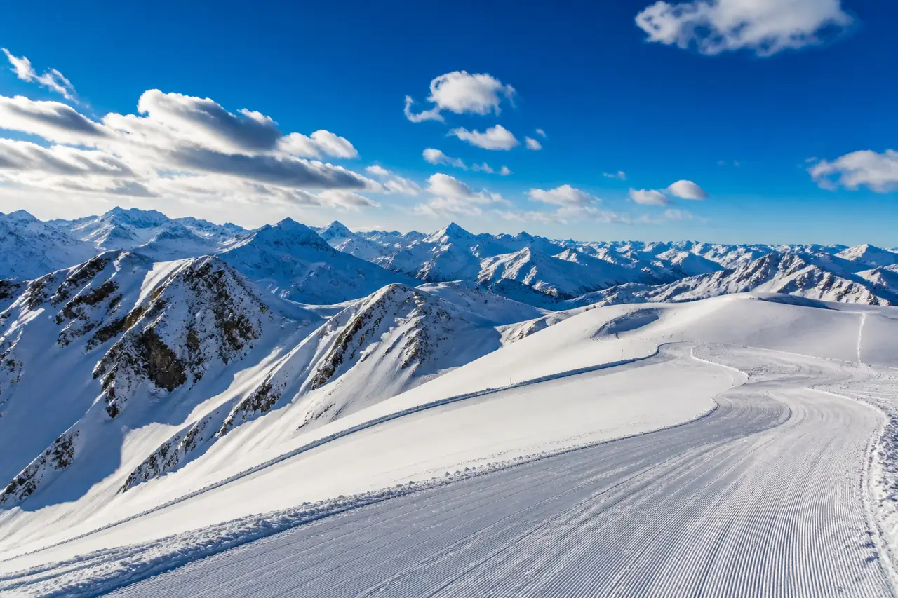 Pista larga che scende da una cupola nevosa in alta quota, con catene di cime innevate all'orizzonte